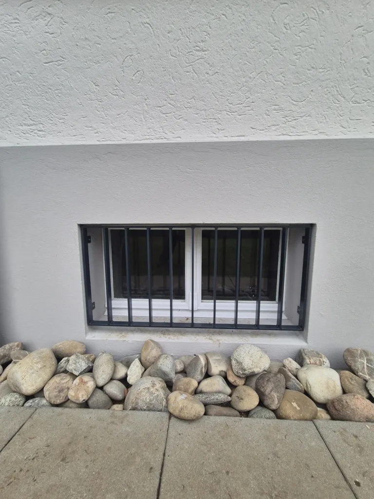 Anthrazitfarbenes Fenstergitter vor einem Kellerfenster in heller Fassade